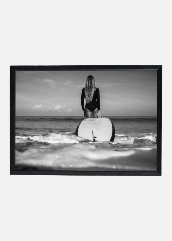 BEAUTIFUL SURFER GIRL IN BLACK BODYSUIT WITH LONG BLOND HAIR STANDING AT BEACH WITH BOARD IN HANDS