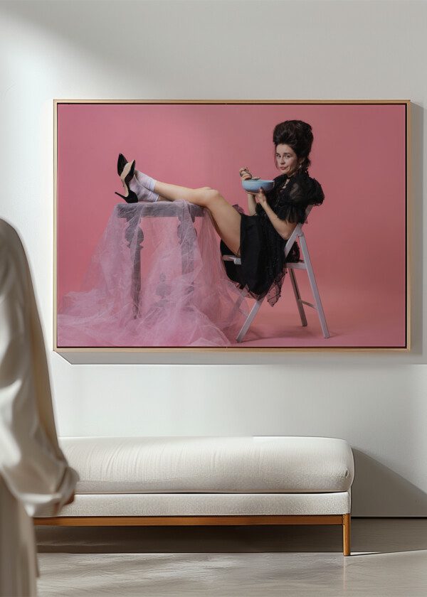 YOUNG PRETTY WOMAN WITH RETRO STYLE HAIRDO IN BLACK EVENING LITTLE DRESS SITTING AT TABLE AND EATING PORRIDGE ISOLATED ON PINK B
