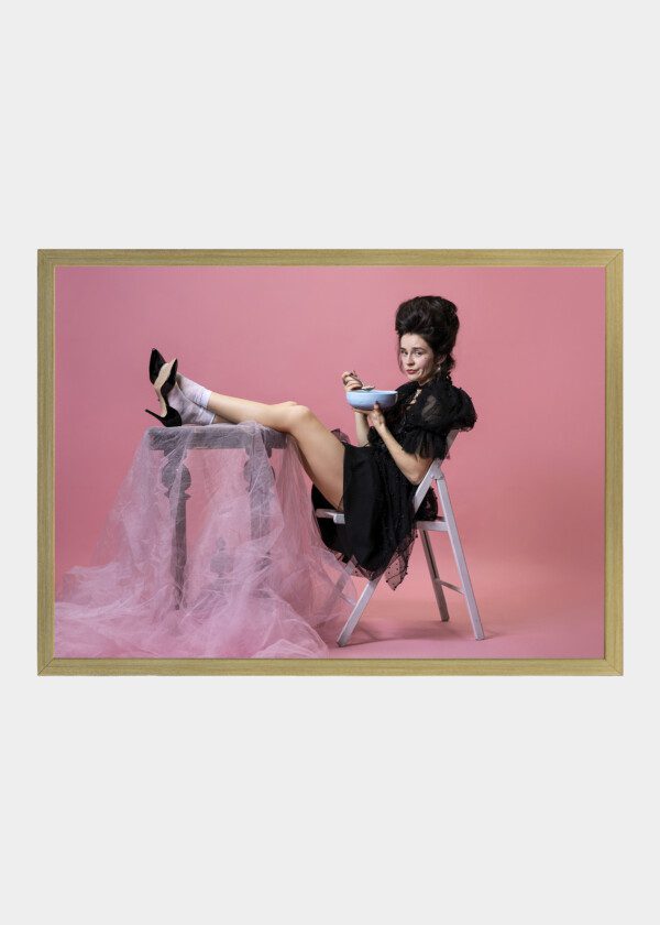 YOUNG PRETTY WOMAN WITH RETRO STYLE HAIRDO IN BLACK EVENING LITTLE DRESS SITTING AT TABLE AND EATING PORRIDGE ISOLATED ON PINK B